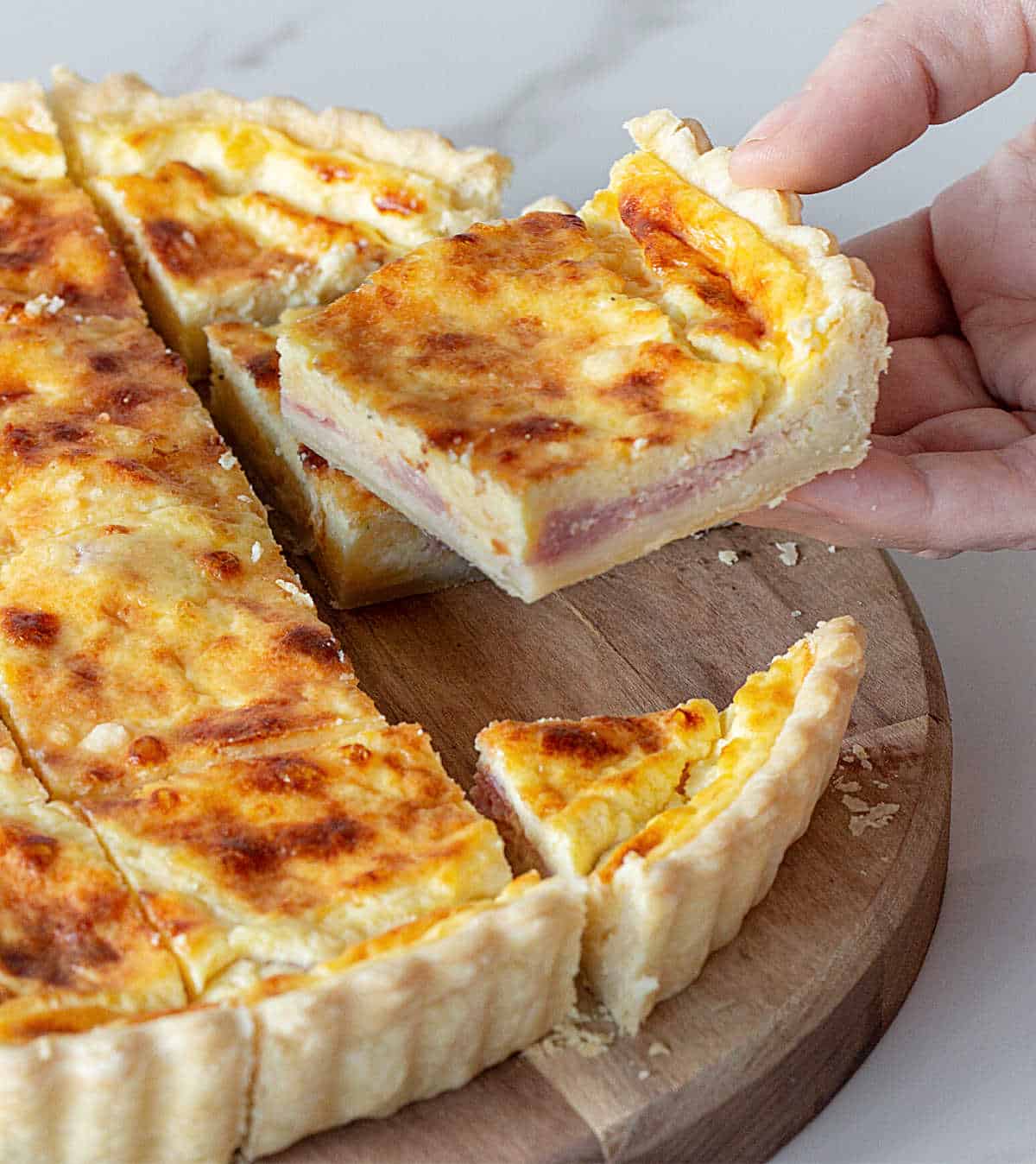 A hand lifting square of ham and cheese quiche on wooden board