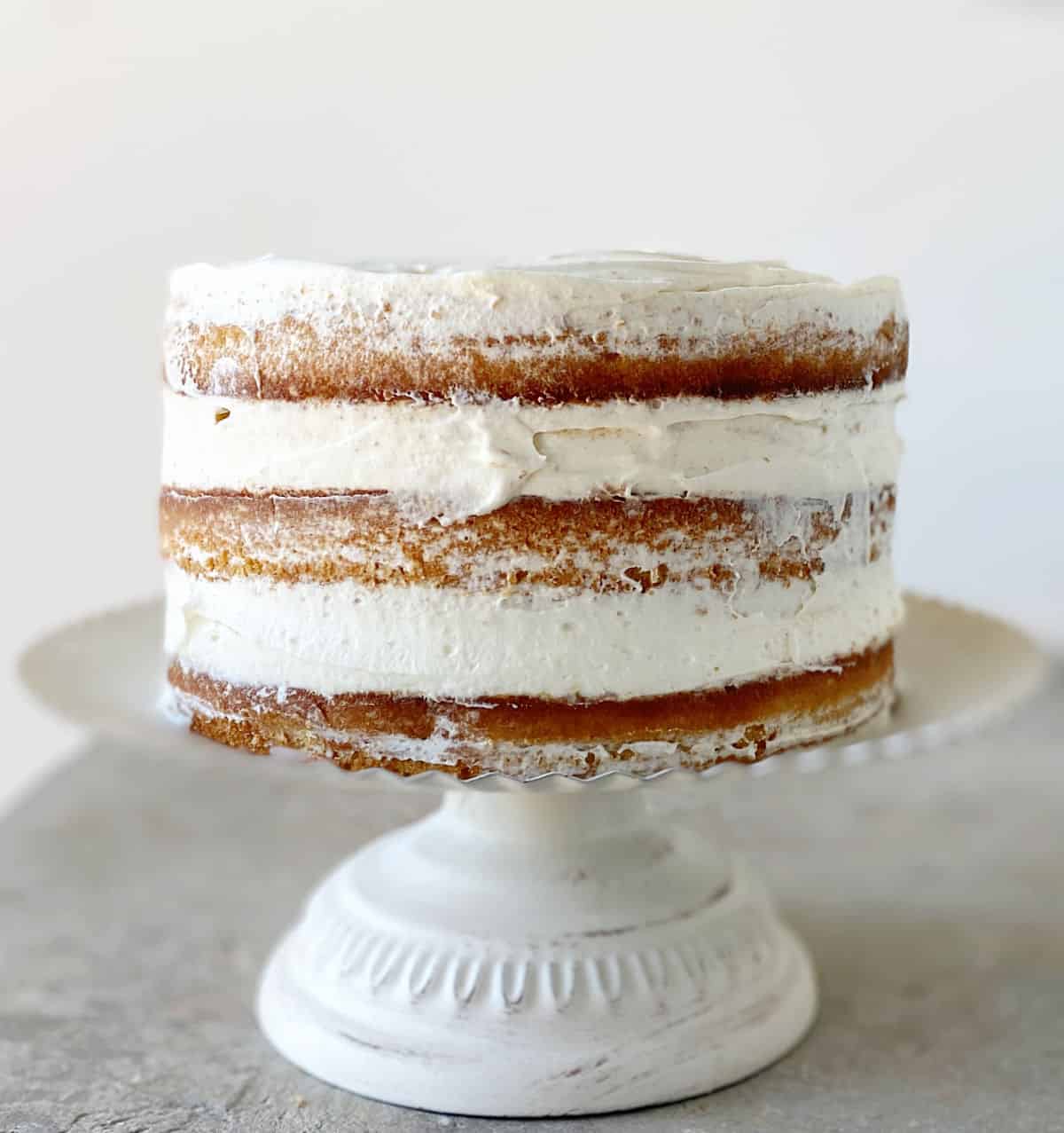 Frosted naked layer cake on white cake stand on grey surface.
