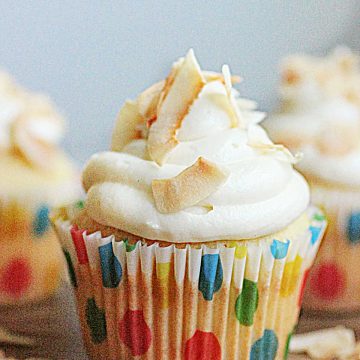 One central white frosted cupcake in dotted paper liner on wooden table with coconut flakes