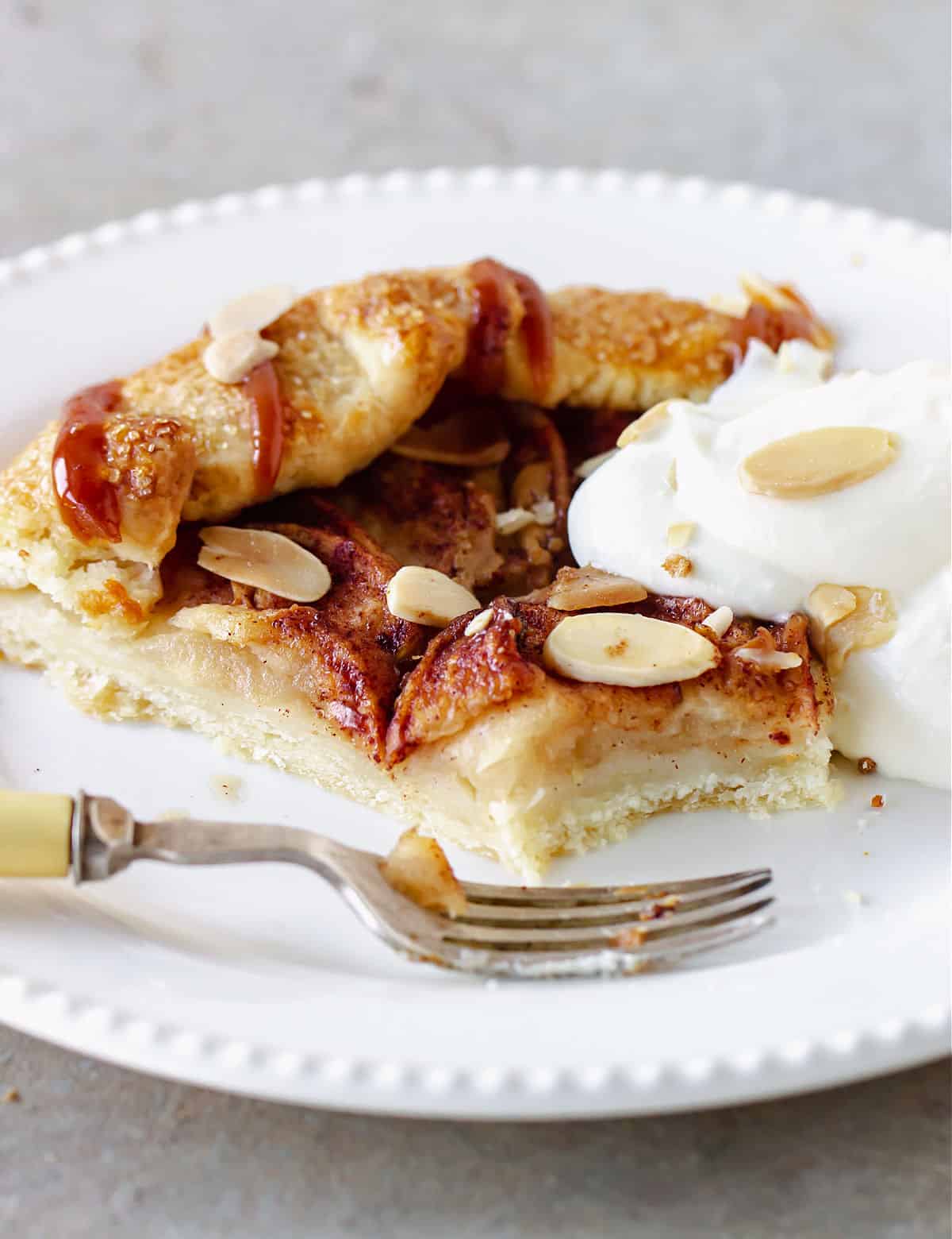 Slice of eaten apple galette with dollop of whipped cream on white plate with fork, grey surface