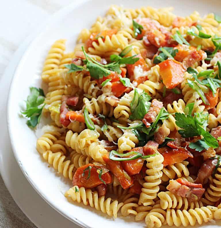 White plate with fusilli pasta, carrots, bacon and chopped parsley