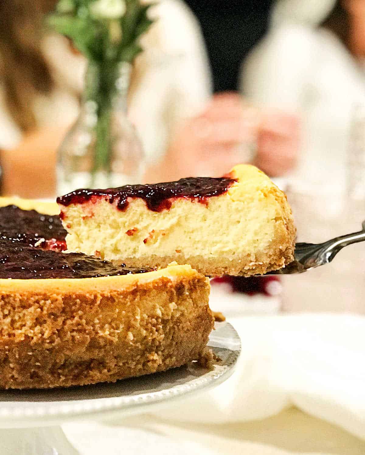 Round cheesecake in table with people in background, a slice being lifted with a cake server