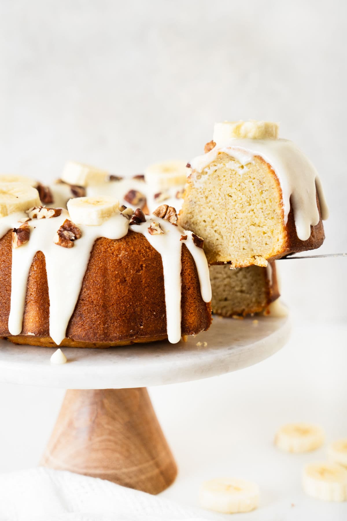 Lifting a slice of glazed banana bundt cake on a wood and marble cake stand. White surface and background. 