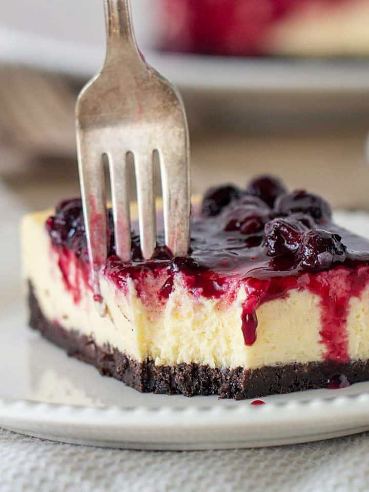 Close-up of cheesecake square on white plate, fork digging into it, berry topping