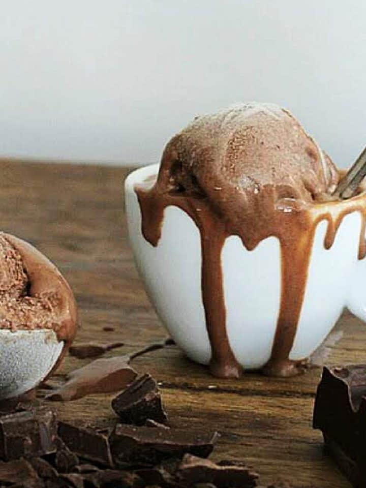 Scoop of chocolate ice cream in a white cup on wooden table, chopped chocolate around it