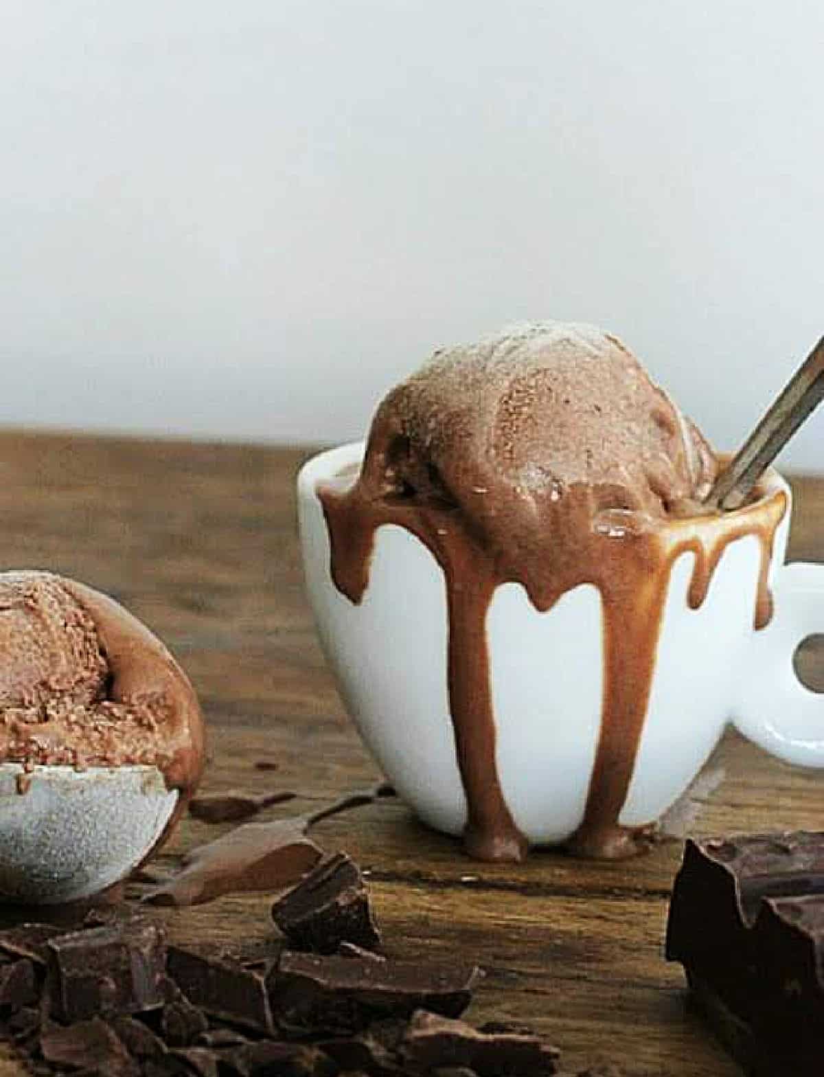 White cup with slightly melting chocolate ice cream on a wooden table, chocolate pieces around