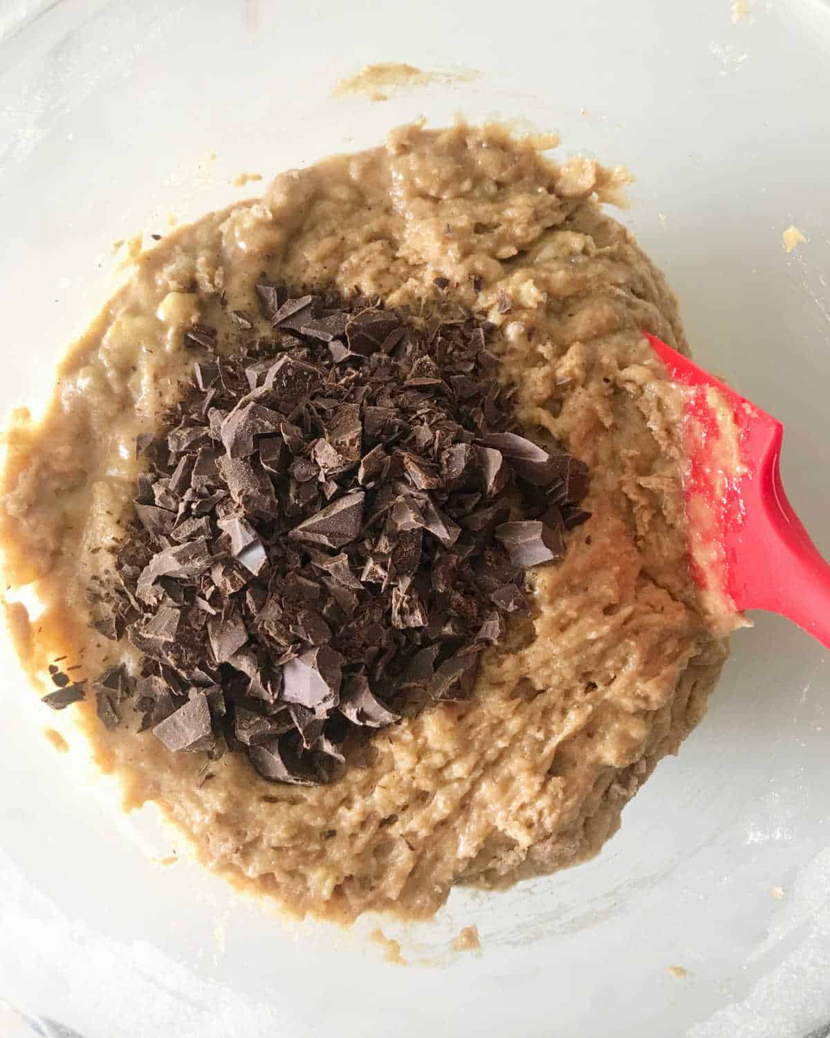 Chocolate chunks added to banana bread batter in a glass bowl with a red spatula.