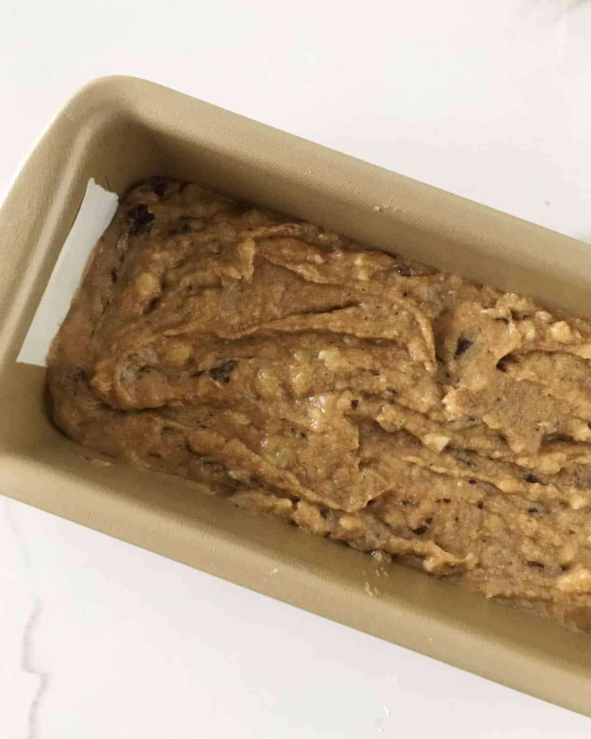 Partial view of banana bread batter in a golden loaf pan on a white surface.