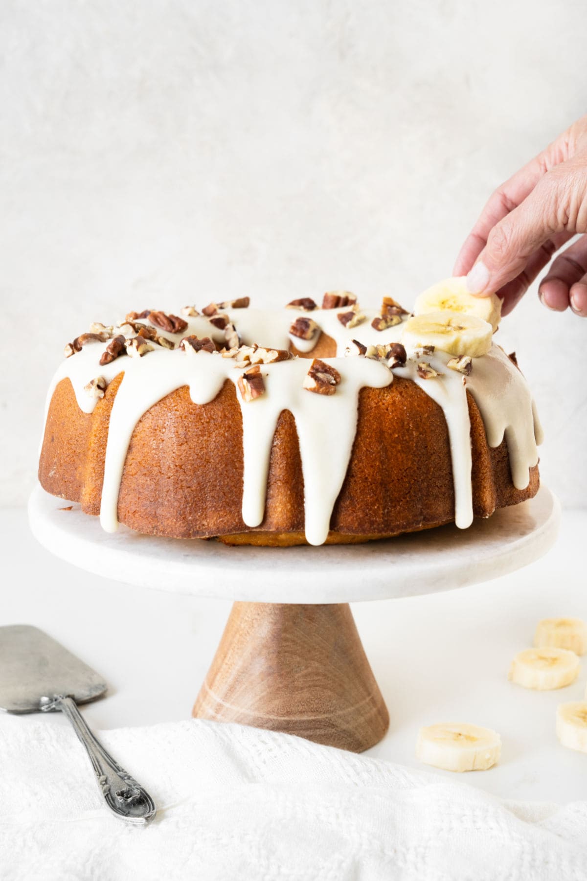 Placing banana rounds and pecans on top of glazed bundt cake on a cake stand. White background. 