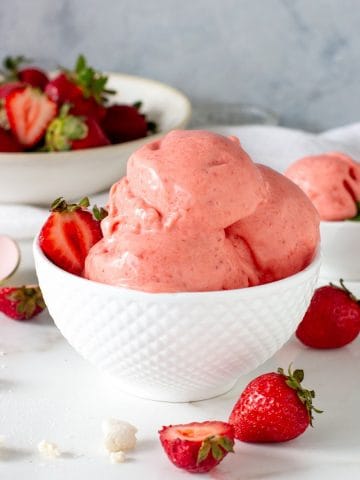 Scoops of strawberry ice cream in white bowl, more strawberries around, white surface, grey background.