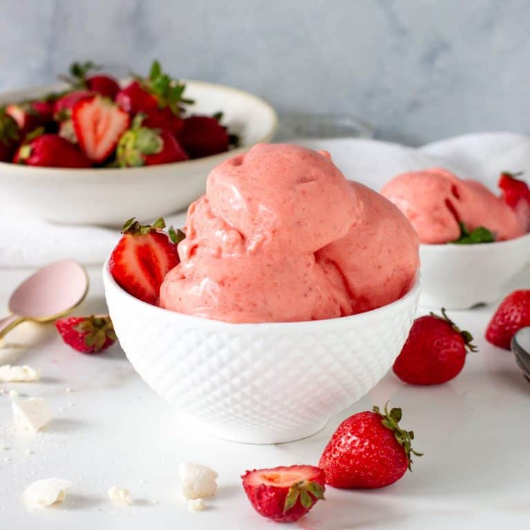 Scoops of strawberry ice cream in white bowl, more strawberries around, white surface, grey background.