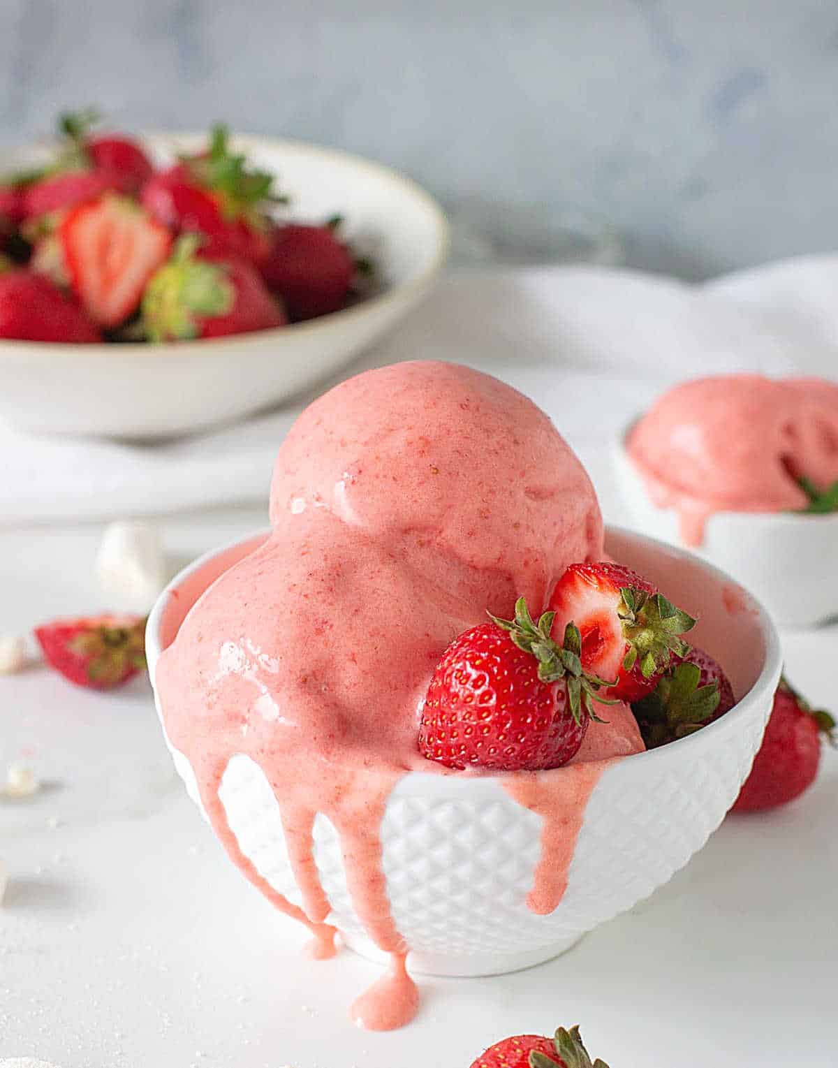 Dripping strawberry ice cream scoops in a white bowl, scattered berries, white surface, grey background.