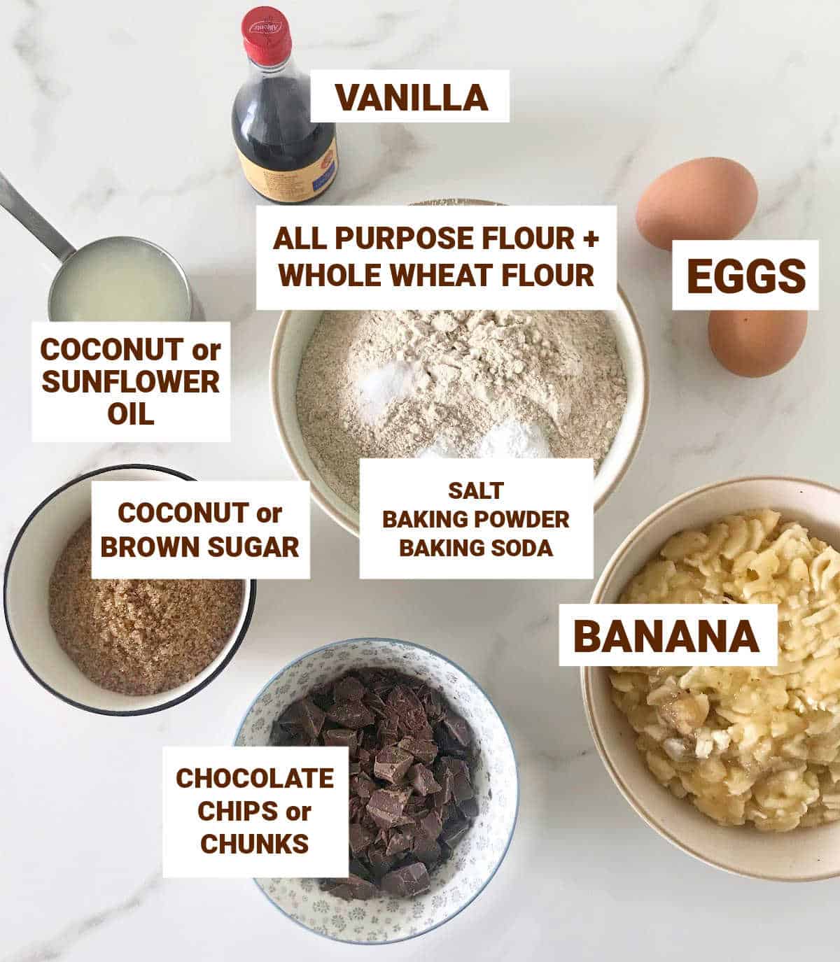 Banana chocolate bread ingredients in bowls on white surface, including eggs, vanilla and chocolate chunks.