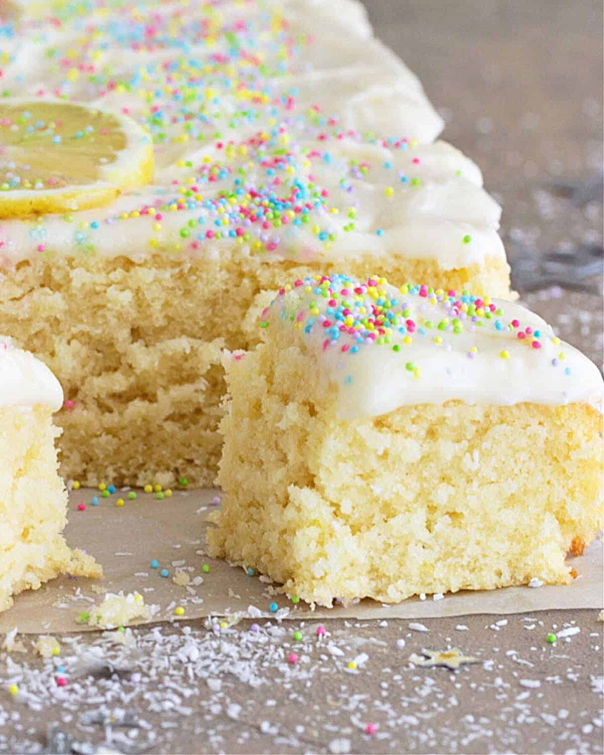 Close up of whole and cut squares of coconut cake with frosting and confetti on grey surface