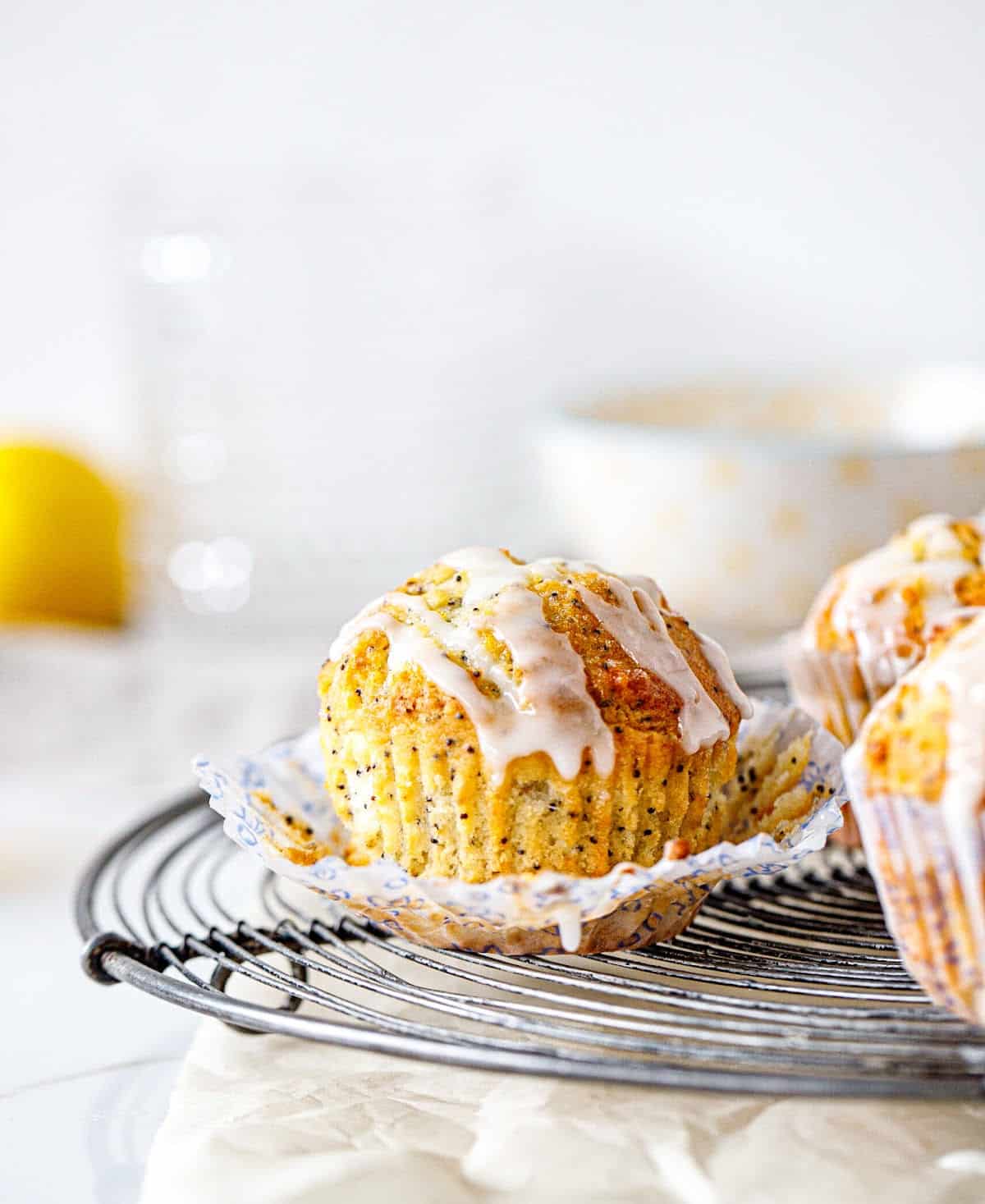 Open paper liner with glazed lemon muffin on a round wire rack with white background.