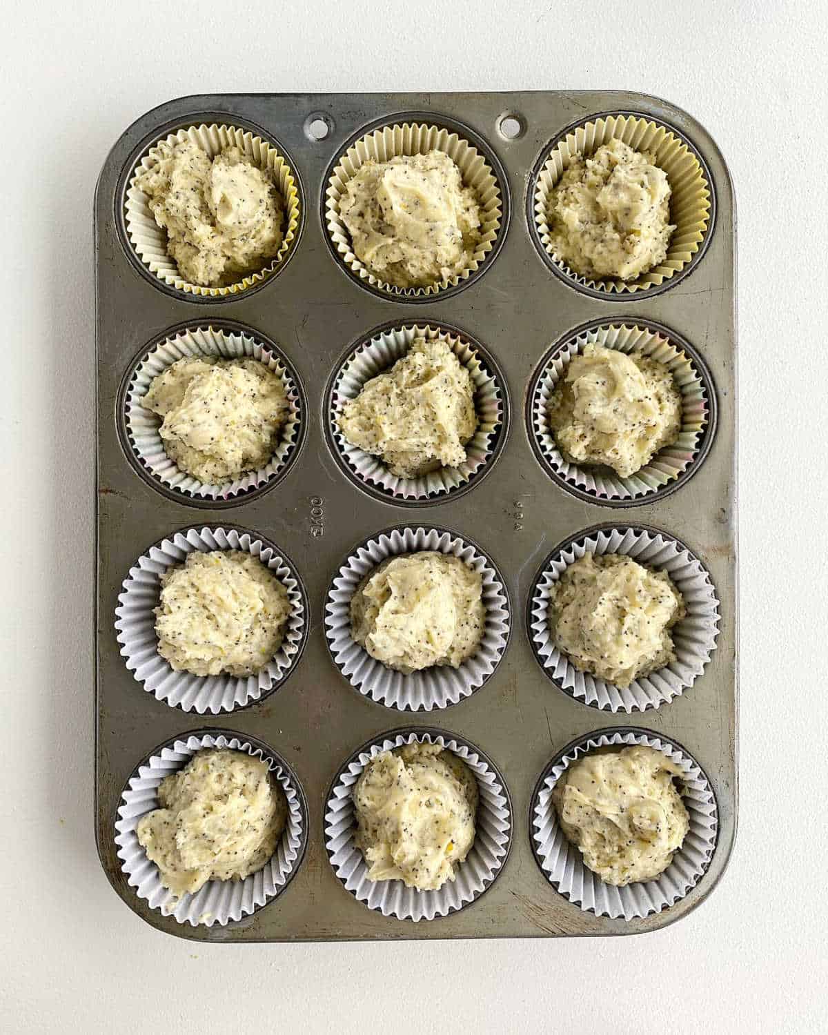 Metal muffin pan filled with lemon poppy seed batter on a white surface.