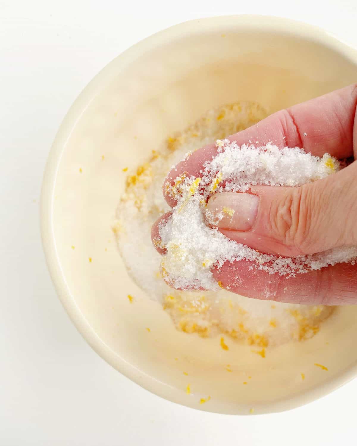 Hand rubbing lemon zest into granulated sugar in a  light beige bowl. 