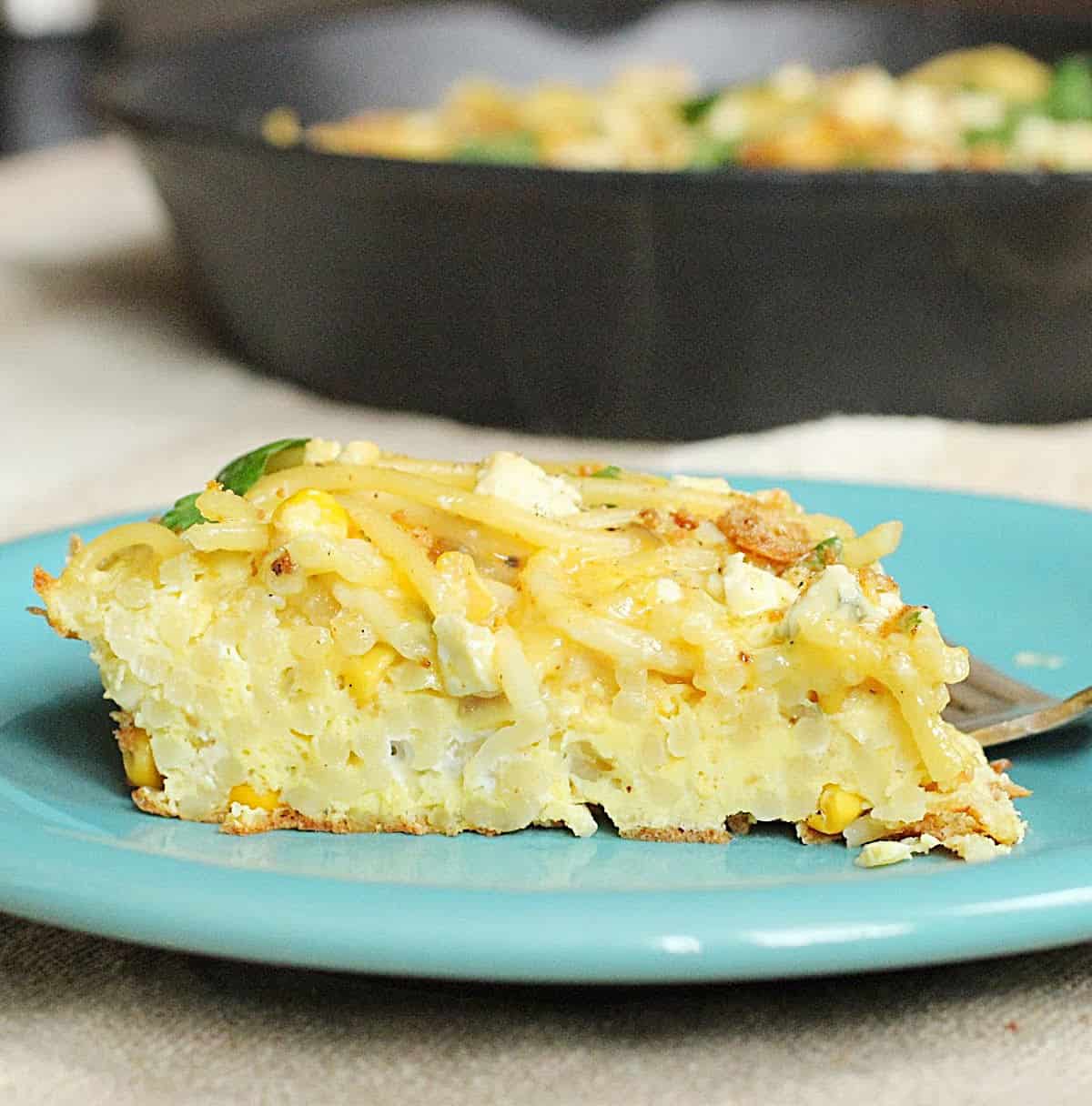 Slice of pasta frittata on a light blue plate with cast iron skillet in the background.