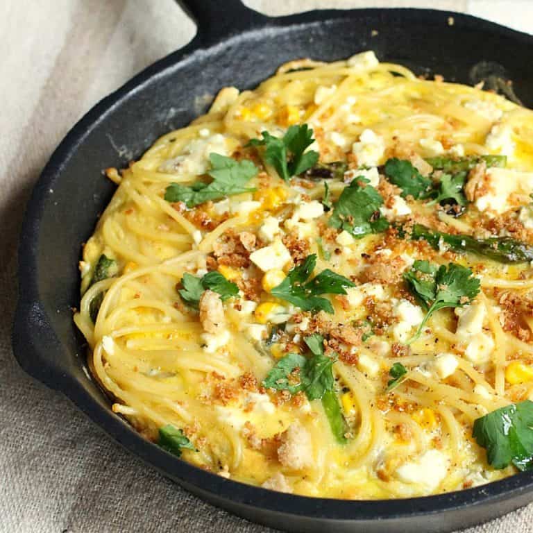 Close up of baked pasta frittata with parsley in a cast iron skillet on a beige surface.