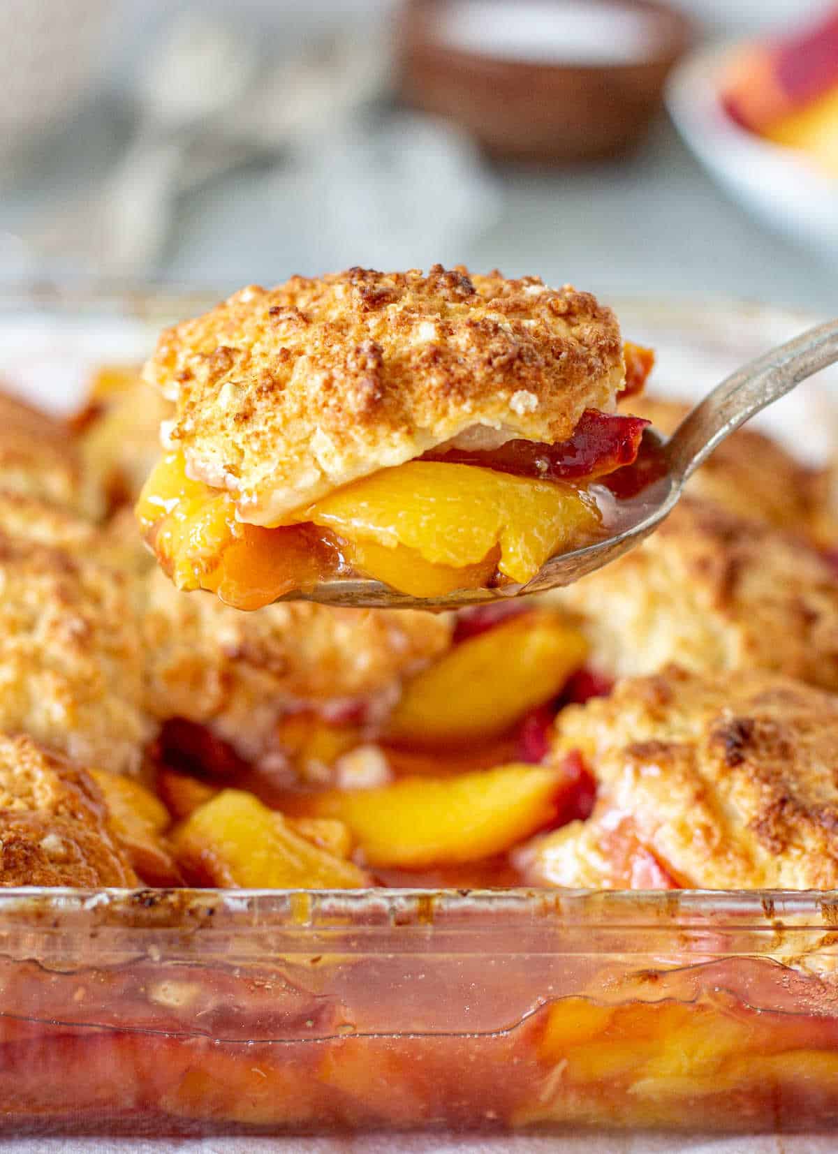 Lifting portion of peach cobbler from glass dish with silver spoon.