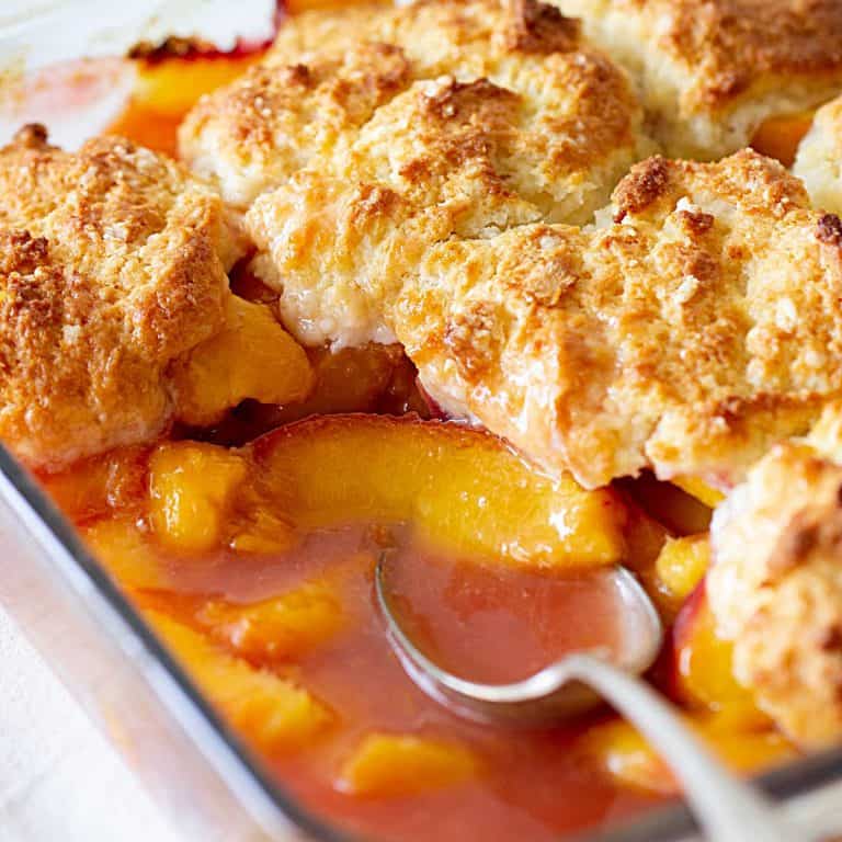 Glass dish with half eaten peaches in syrup, biscuit topping and silver spoon.