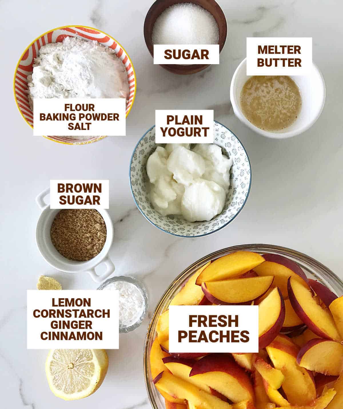 White surface with bowls containing ingredients for peach cobbler including yogurt, butter, lemon, and brown sugar.