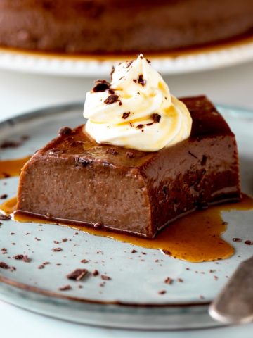 Cream topped serving of chocolate flan on grey blue plate with silver spoon
