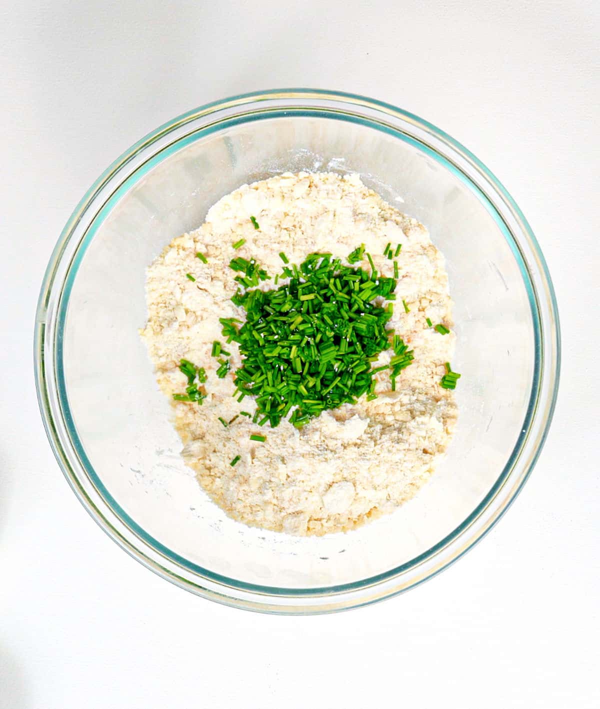 Glass bowl with scone ingredients and chopped chives. White surface.