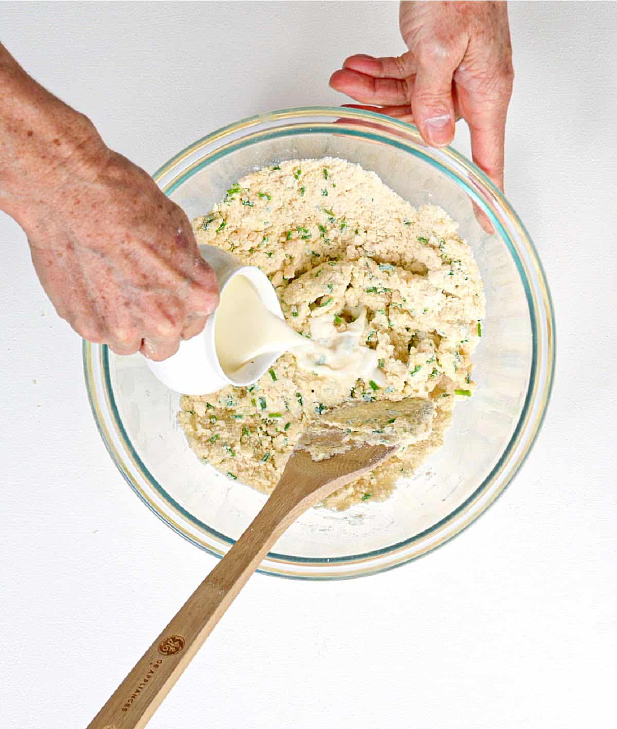 Adding milk to a glass bowl with cheese chive scone dough ingredients. White surface.