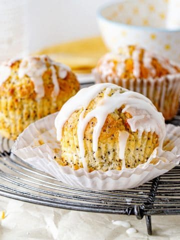 Several glazed lemon poppy seed muffins in paper liners on a wire rack. White and gray background.