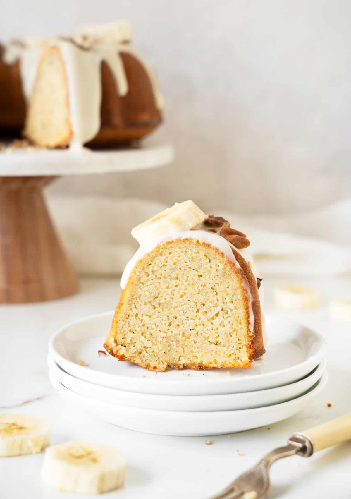 Stack of white plates with slice of glazed banana bundt cake. White surface and rest of cake in the background. 