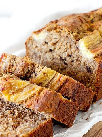 White parchment paper with loaf of banana bread with some cut slices, white background.