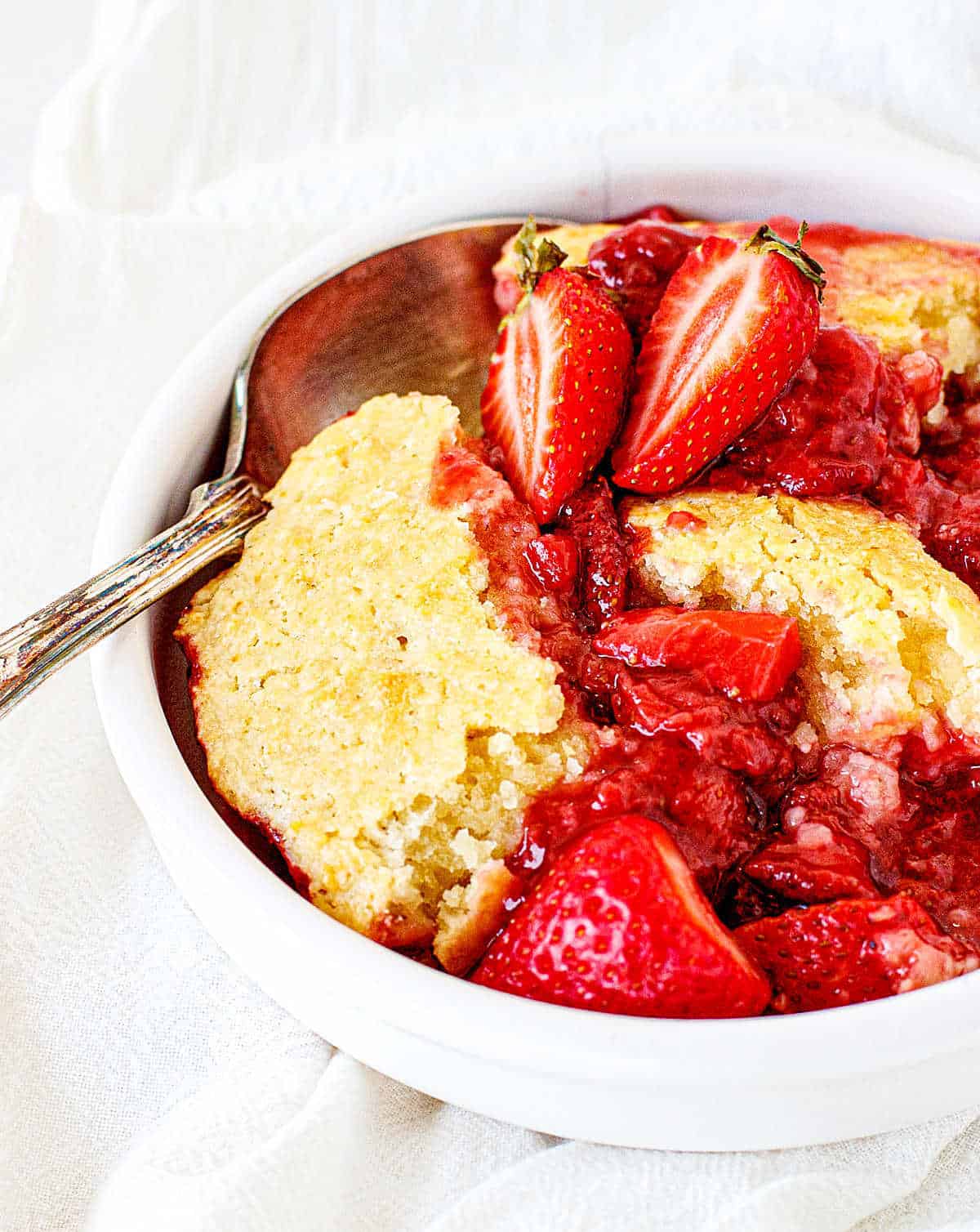 White bowl with strawberry cobbler serving, silver spoon, white background