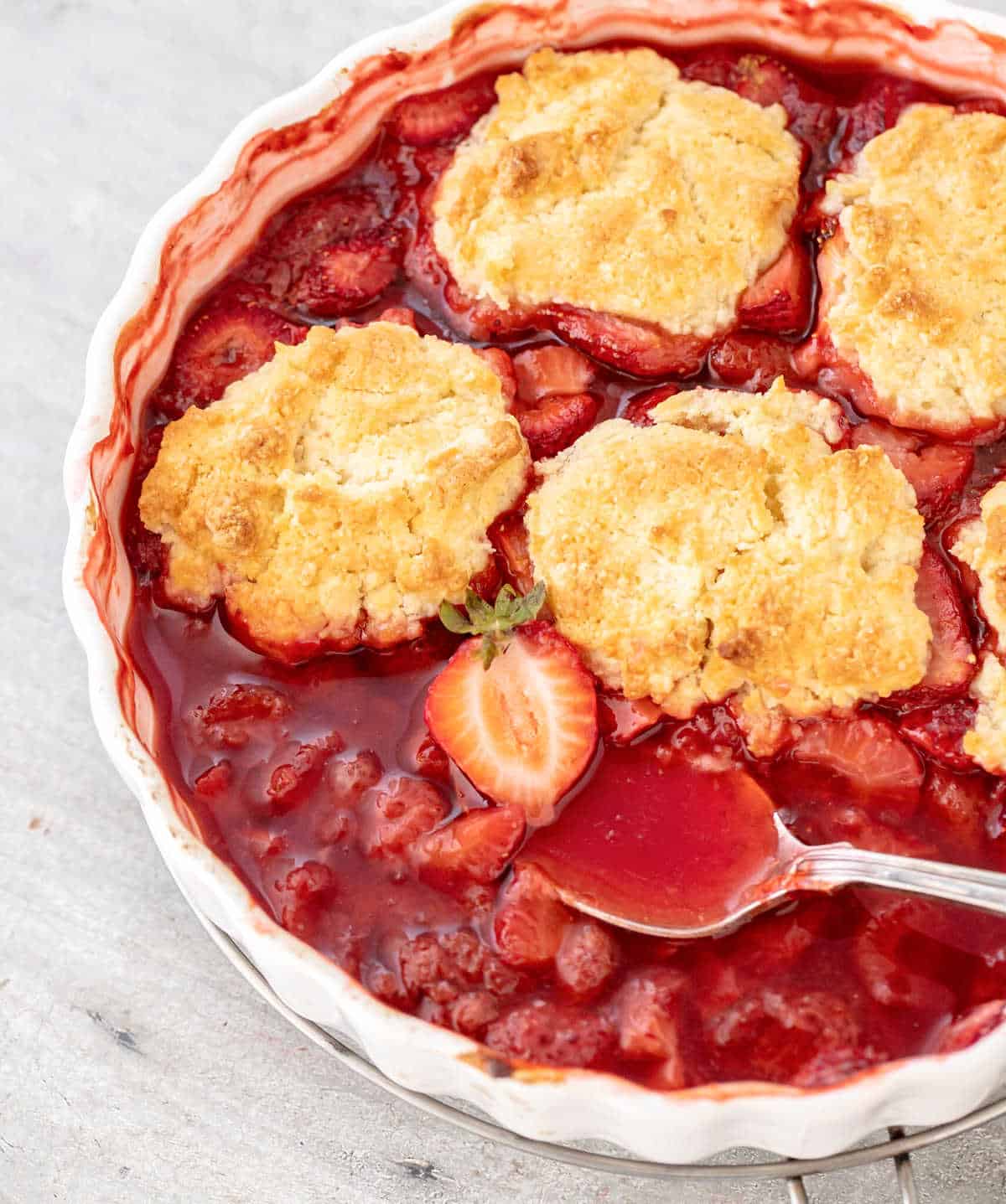 Close up of strawberry cobbler with silver spoon in white dish on grey surface.