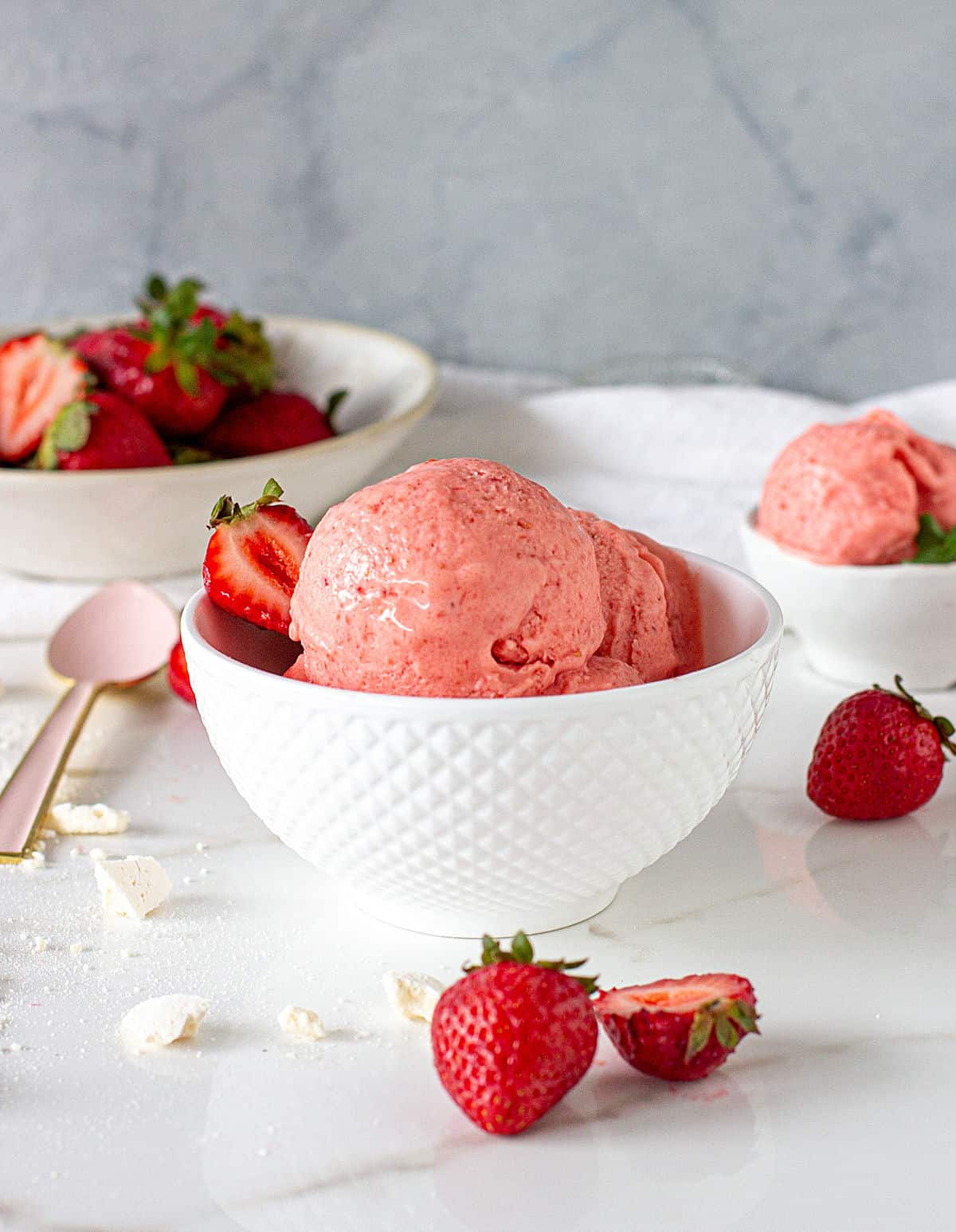 White bowls with strawberry ice cream and whole strawberries, scattered strawberries, grey background
