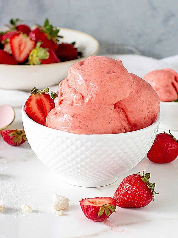 Scoops of strawberry ice cream in white bowl, more strawberries around, white surface, grey background