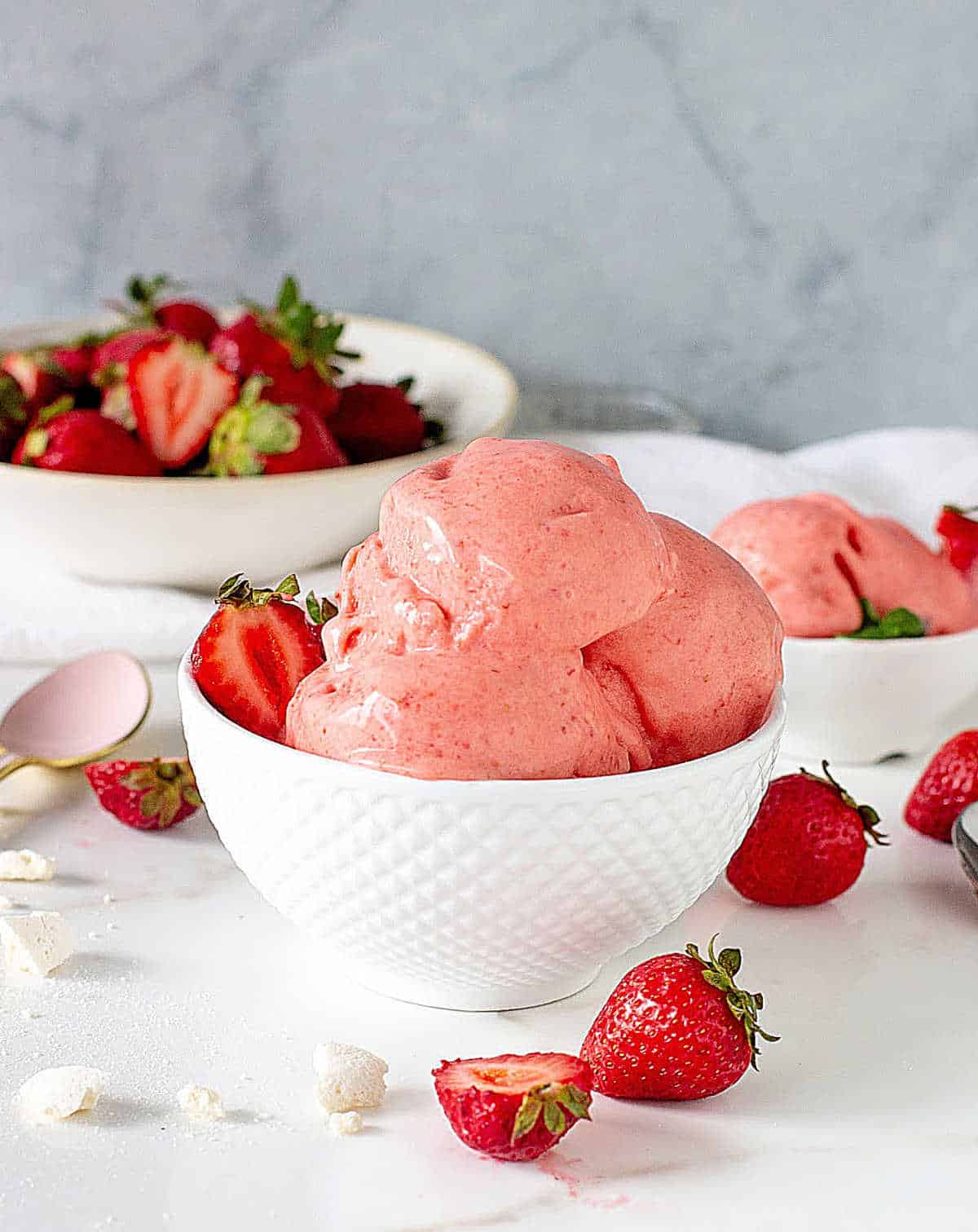 Several scoops of strawberry ice cream in white bowl, more strawberries around, white surface, grey background.