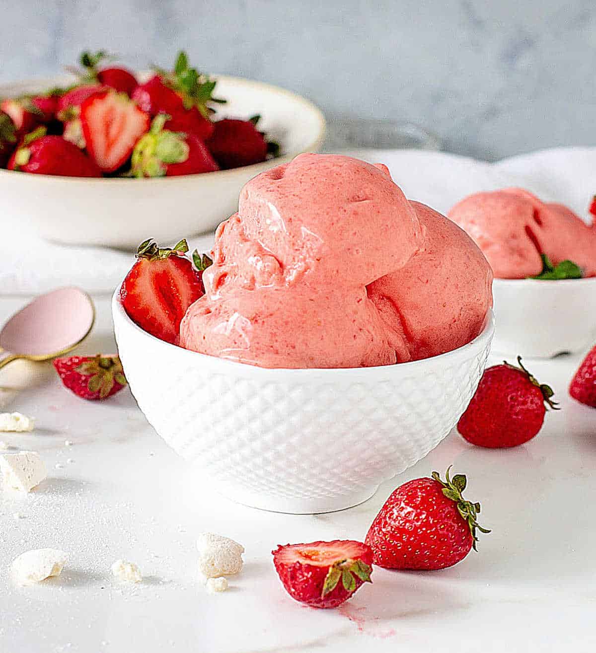 White bowls with scoops of strawberry ice cream. White marble surface, grey background, and fresh strawberries around. 