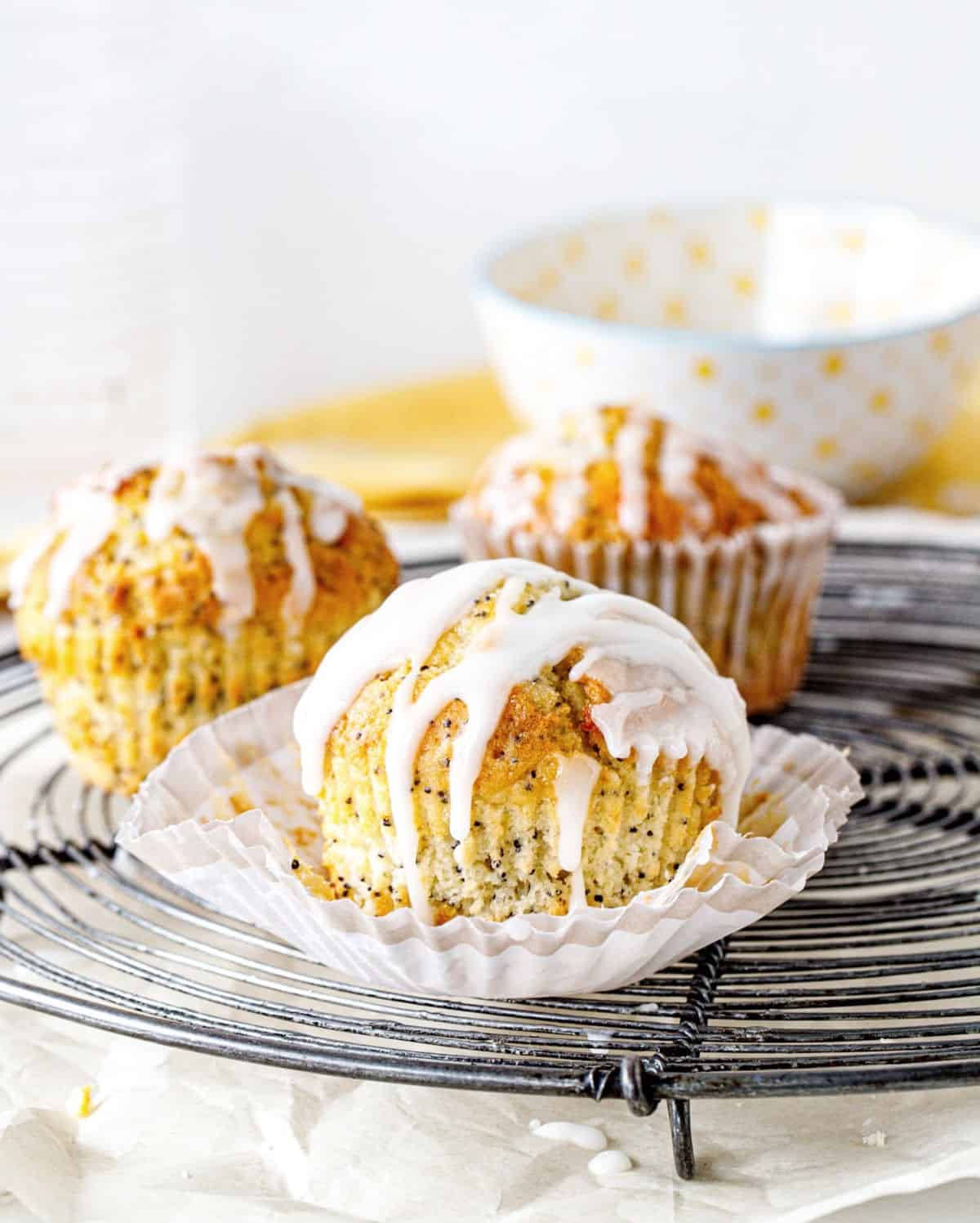 Three glazed lemon poppy muffins on a metal round cooling rack; bowl in the white background.
