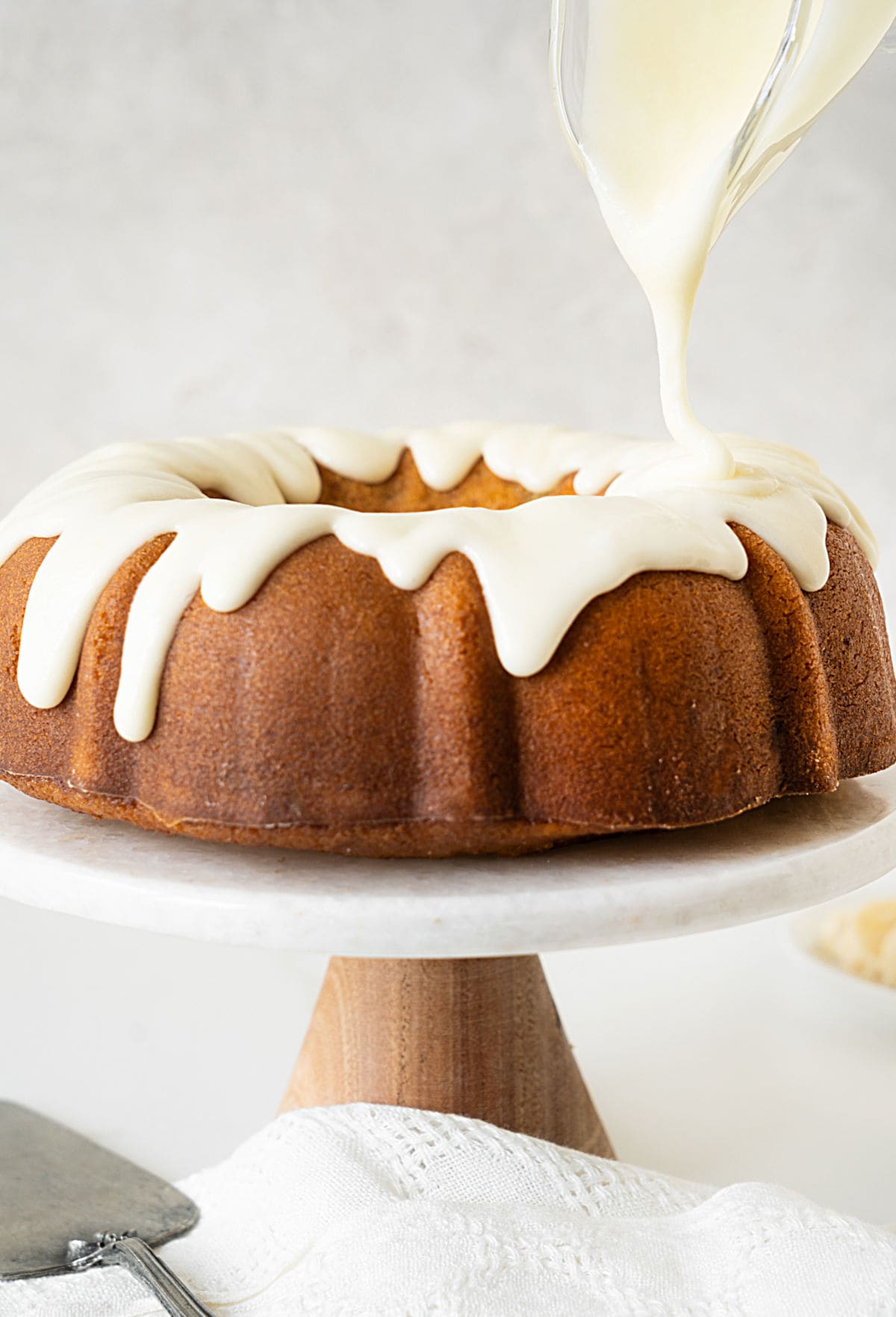 Drizzling a bundt cake on a cake stand with cream cheese glaze. Light grey background.