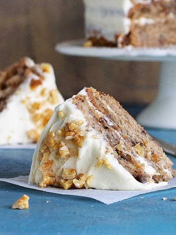 Blue surface with slice of hummingbird cake, more in brown background on cake stand