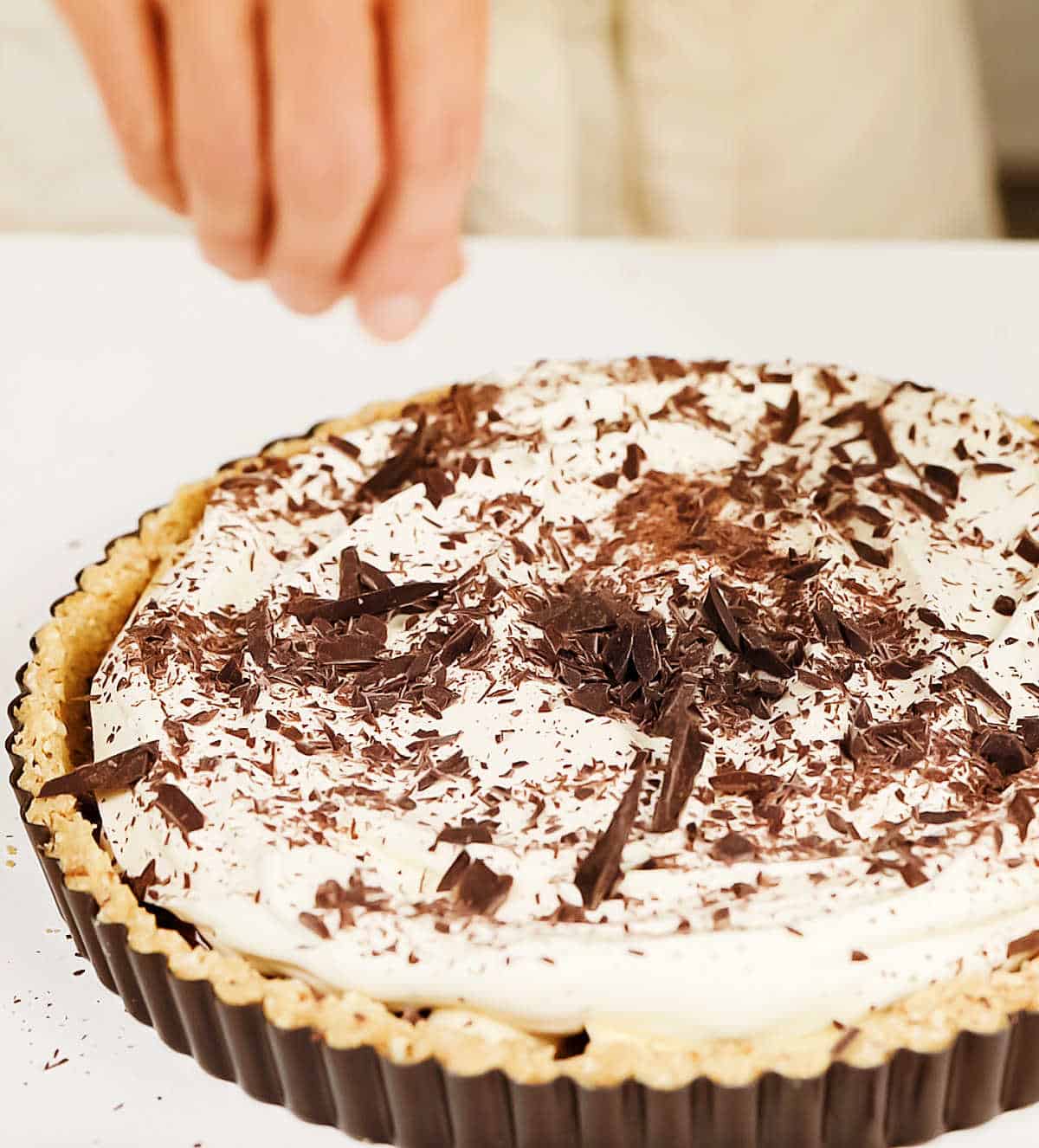 Hand sprinkling grated chocolate on whipped cream in pie pan on white surface