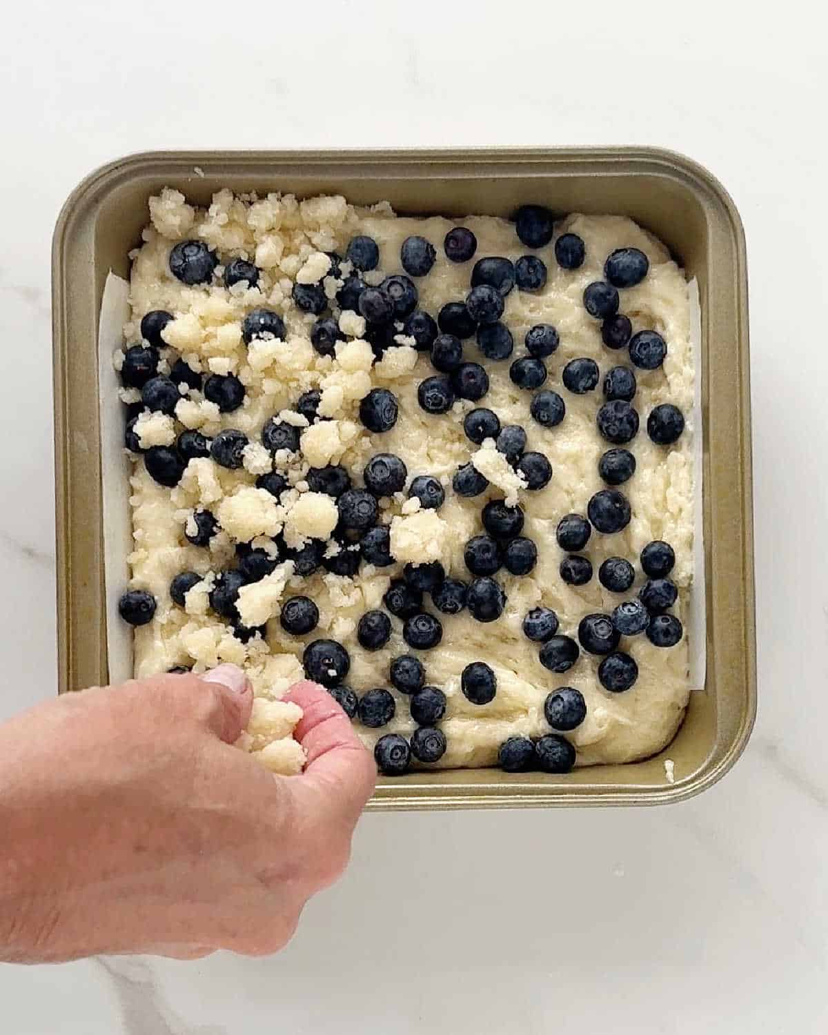Scattering crumble topping to a blueberry cake in a square metal pan on white marble.