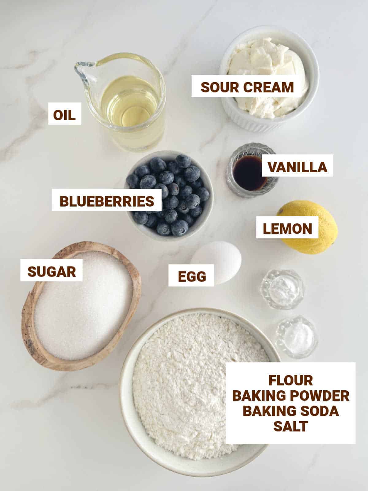 Bowls on a white surface containing ingredients for blueberry cake including egg, lemon, sugar, oil, vanilla, flour, sour cream.
