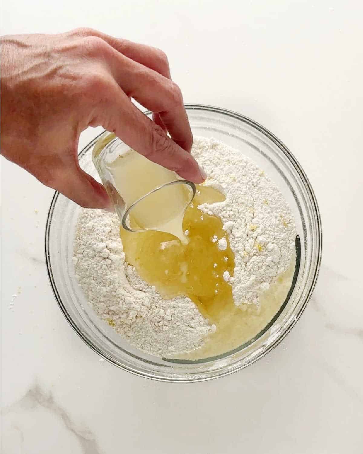Adding lemon juice to glass bowl with flour mixture and oil. White marble surface.