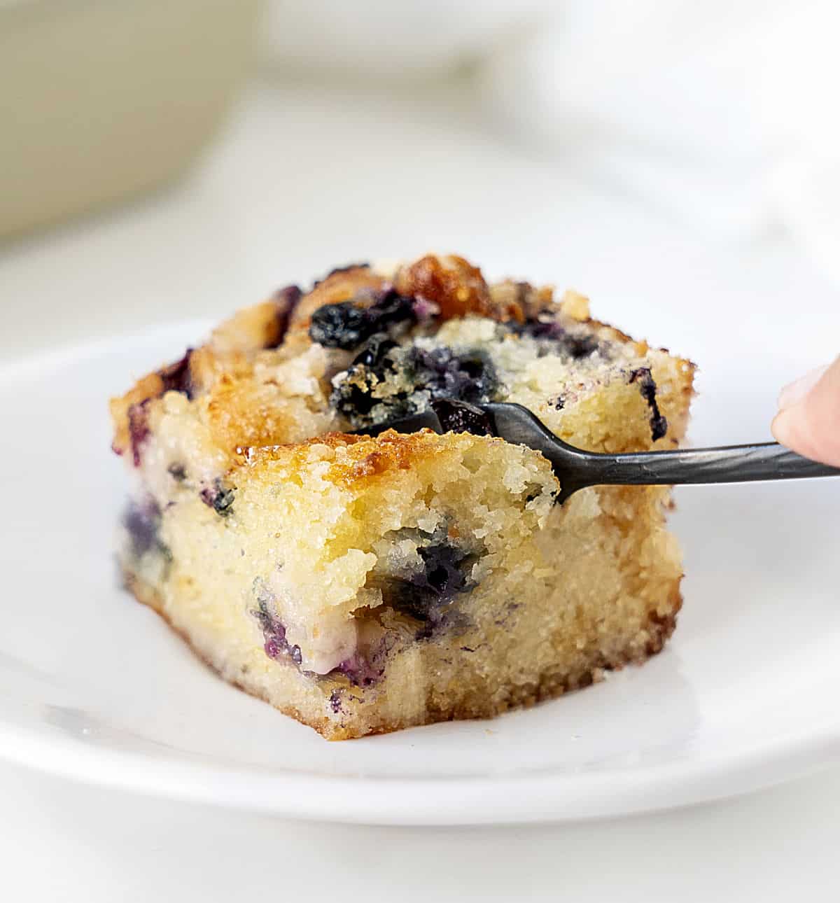 Black fork cutting a square of blueberry crumb cake in a white plate.