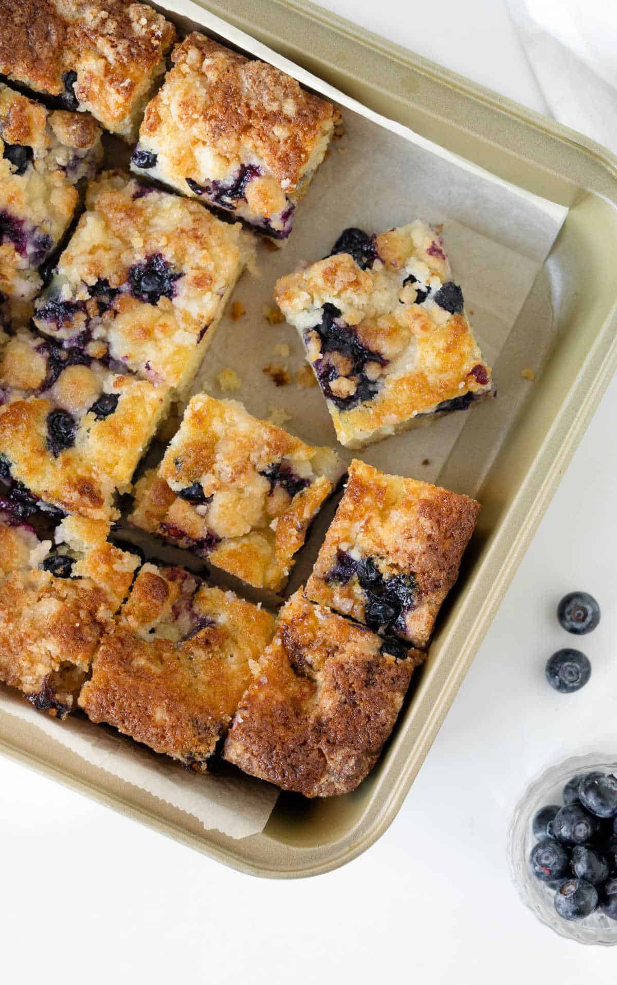 White marble surface with golden metal square pan with blueberry cake squares. 