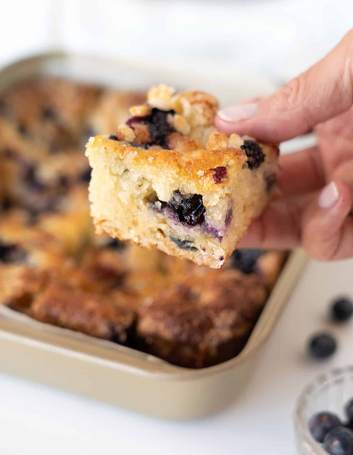 Holding a square of blueberry crumble cake with metal pan and cake in the white background.