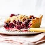 A slice of cherry crumb pie on white plate on pink napkin, yellow fork, white background