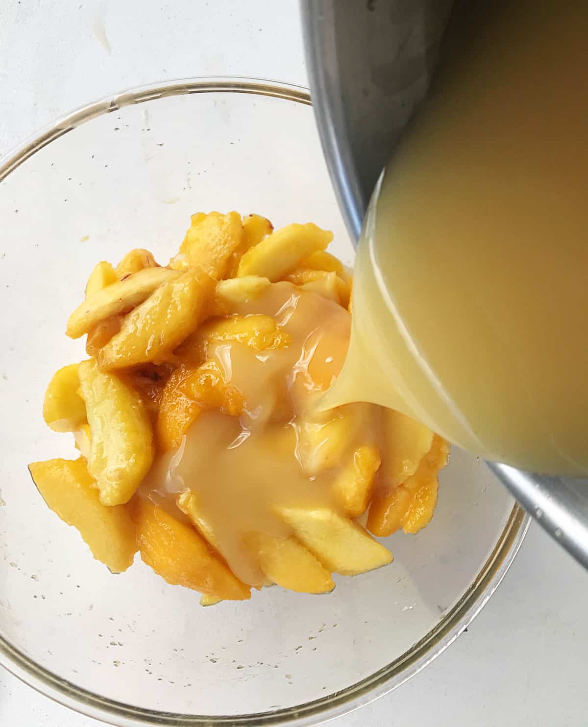 Syrup is added to fresh peach slices in glass bowl on white surface